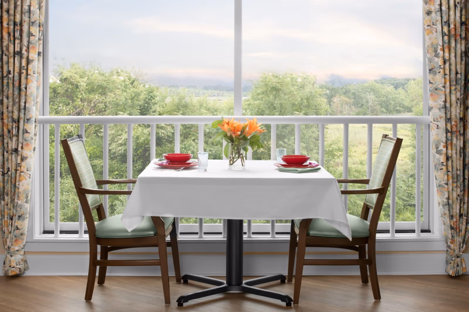 A small dining table set for two with a white tablecloth, red bowls, plates, glasses, and a vase with orange flowers. The table is positioned in front of a large window with a view of green trees and a cloudy sky. Floral curtains frame the window.