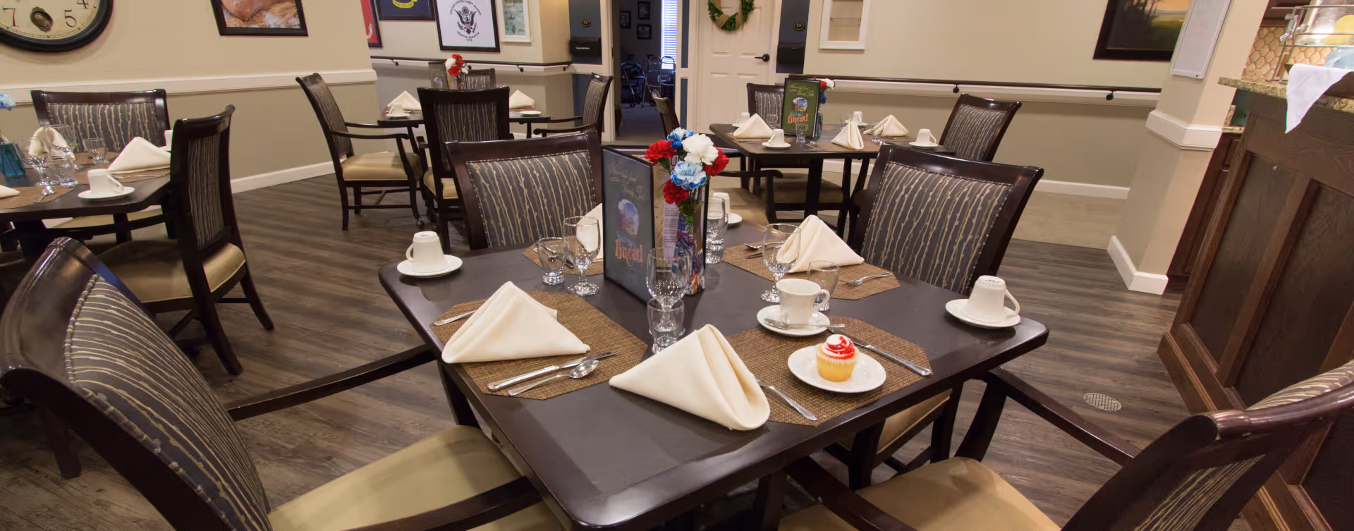 A dining room in a senior living facility with several tables set for a meal. Each table has neatly folded white napkins, cups, glasses, silverware, and a small vase with red, white, and blue flowers. One table has a small cupcake on a plate. The room has wood flooring, beige walls, and framed pictures on the walls.