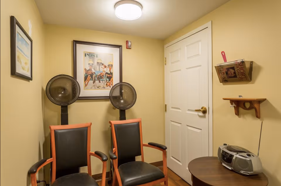 Small room with two black and wooden chairs positioned under two vintage hair dryers mounted on stands. The walls are painted yellow and decorated with two framed pictures. A white door with a gold handle is on the right side. A small wooden table holds a silver radio, and a wall-mounted magazine holder contains a magazine.