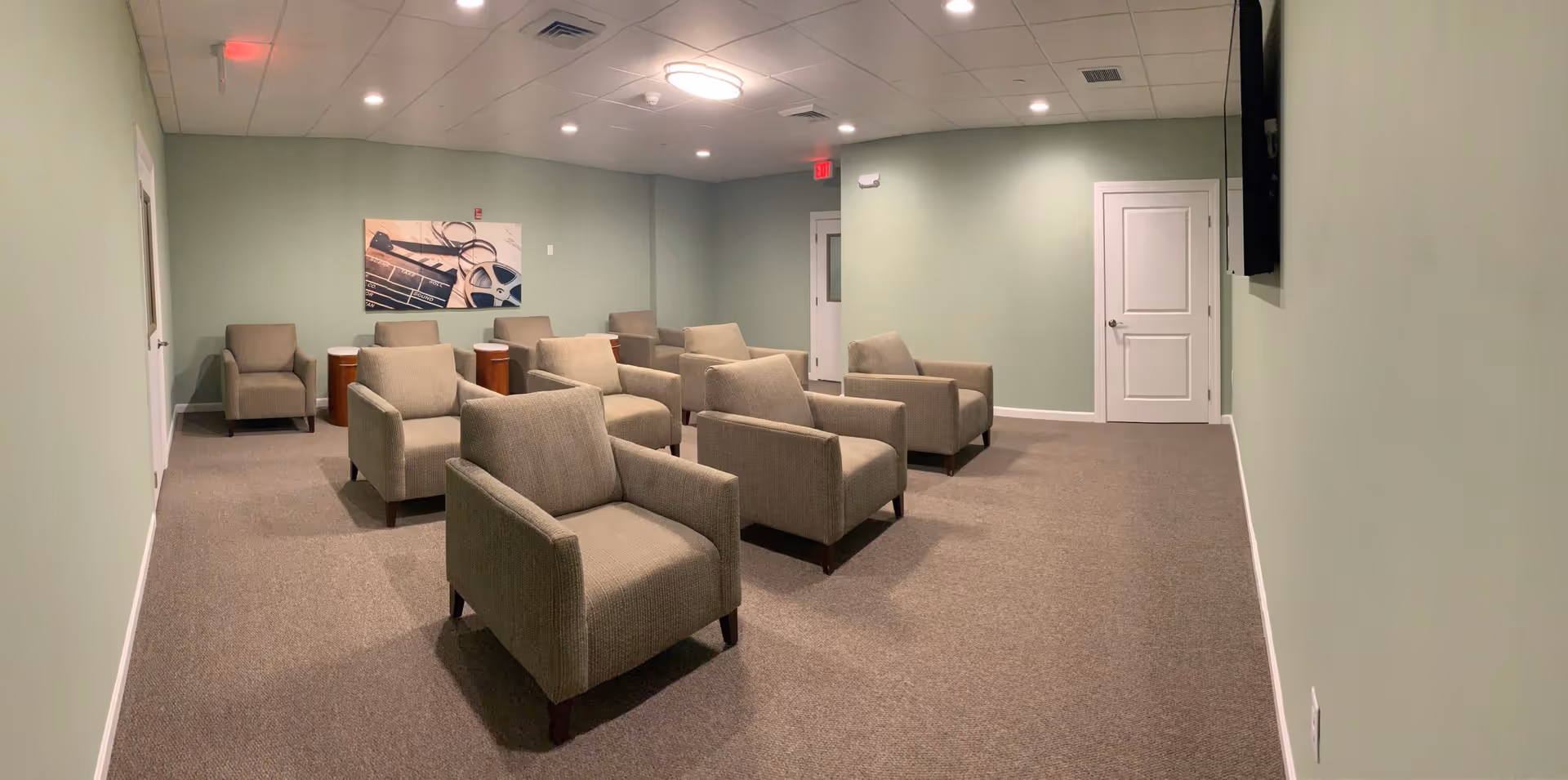 A small theater or media room with two rows of beige armchairs facing a wall-mounted TV. The walls are painted light green, and there is a picture of a film reel and clapperboard on the back wall. The room has a carpeted floor and recessed ceiling lights.