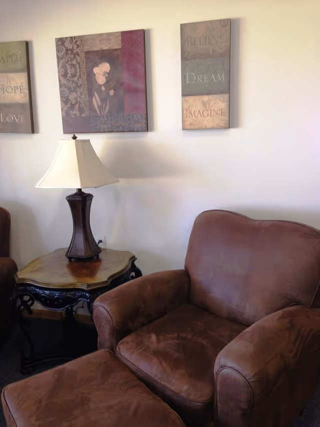 Brown upholstered armchair with matching ottoman next to a wooden side table and lamp beneath framed wall art.