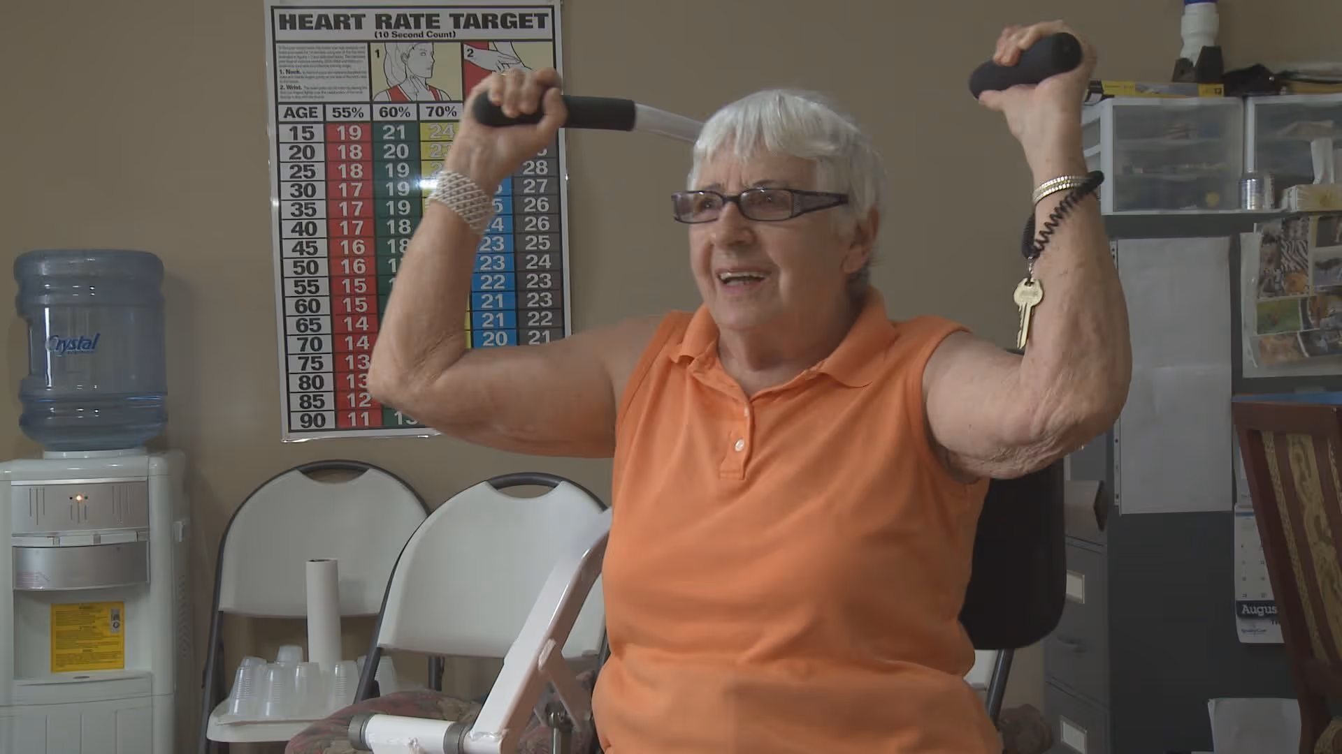 An elderly woman with short white hair and glasses is exercising indoors using a shoulder press machine. She is wearing an orange sleeveless polo shirt and smiling. Behind her, there is a water cooler, two white folding chairs, a heart rate target chart on the wall, and some office furniture with papers and a calendar.