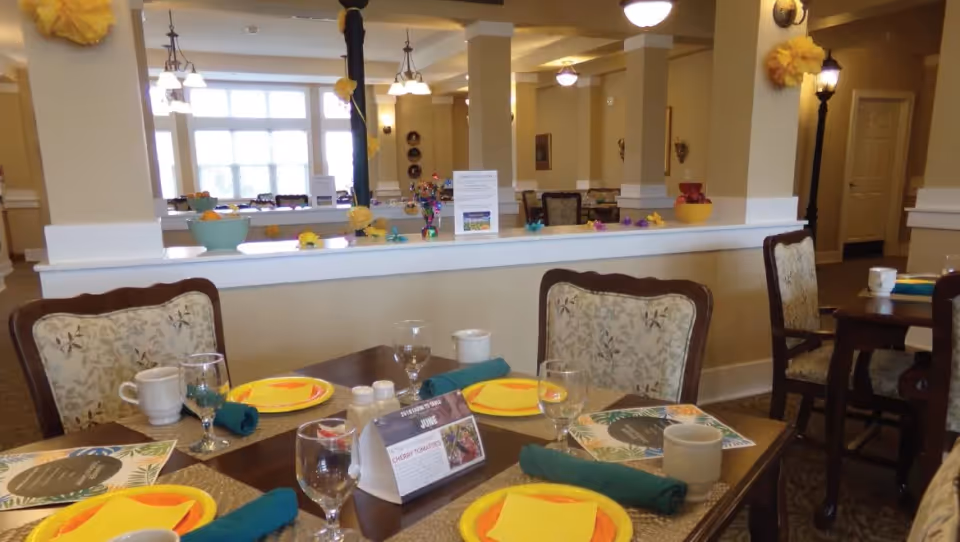 A dining room in a senior living facility with tables set for a meal. The tables have yellow plates, green napkins, glasses, cups, and placemats. The room has beige walls, decorative columns, and large windows letting in natural light. There are chairs with floral upholstery around the tables and decorative items on a half wall in the background.