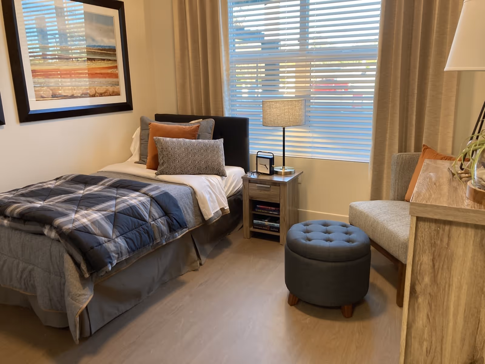 A cozy bedroom with a single bed made with gray and plaid bedding, multiple pillows, a wooden nightstand with a lamp and a small clock, a window with blinds and beige curtains, a gray upholstered ottoman, a cushioned chair with an orange pillow, and a wooden dresser with a plant on top.