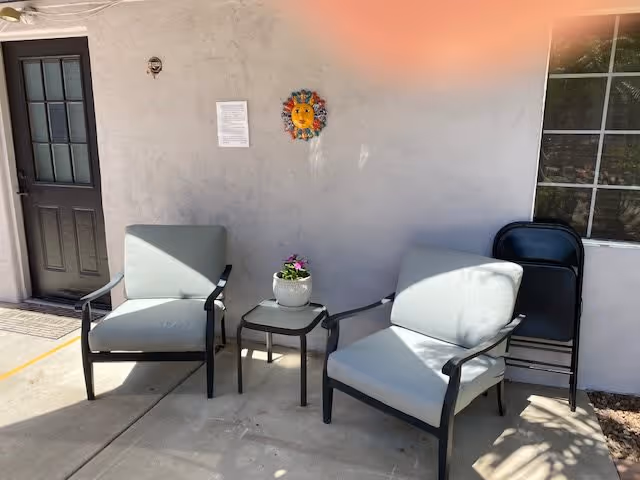 Outdoor patio area with two cushioned armchairs and a small glass-top table between them holding a white pot with purple flowers. The wall behind has a decorative sun face and a small sign. There is a door on the left and a window on the right with two folded black chairs leaning against the wall.