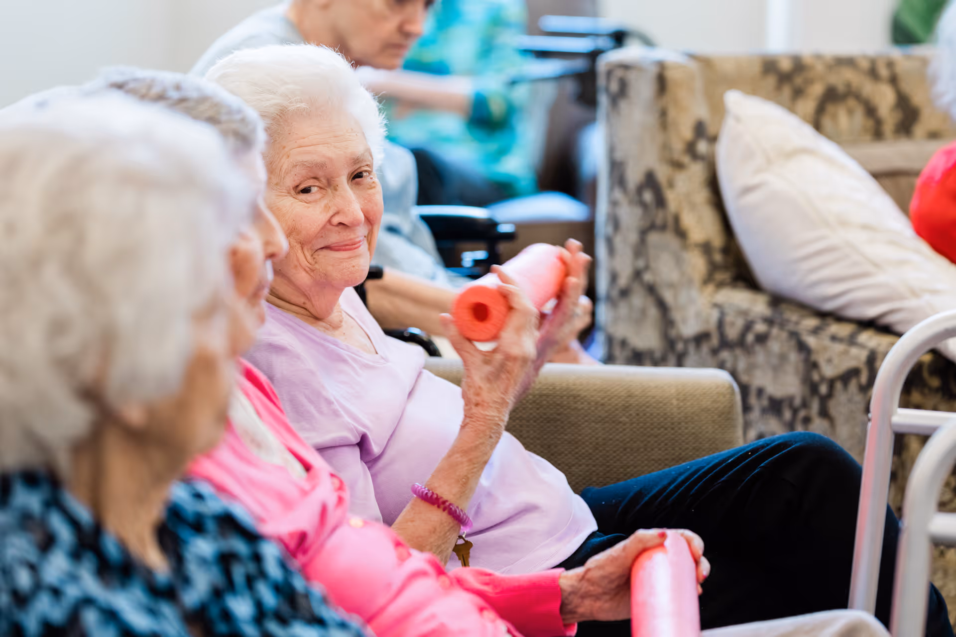 A group of elderly women sitting together in a common area, one woman in a light purple shirt is smiling and holding a red foam exercise roller, with others seated nearby and a patterned couch with a white pillow in the background.