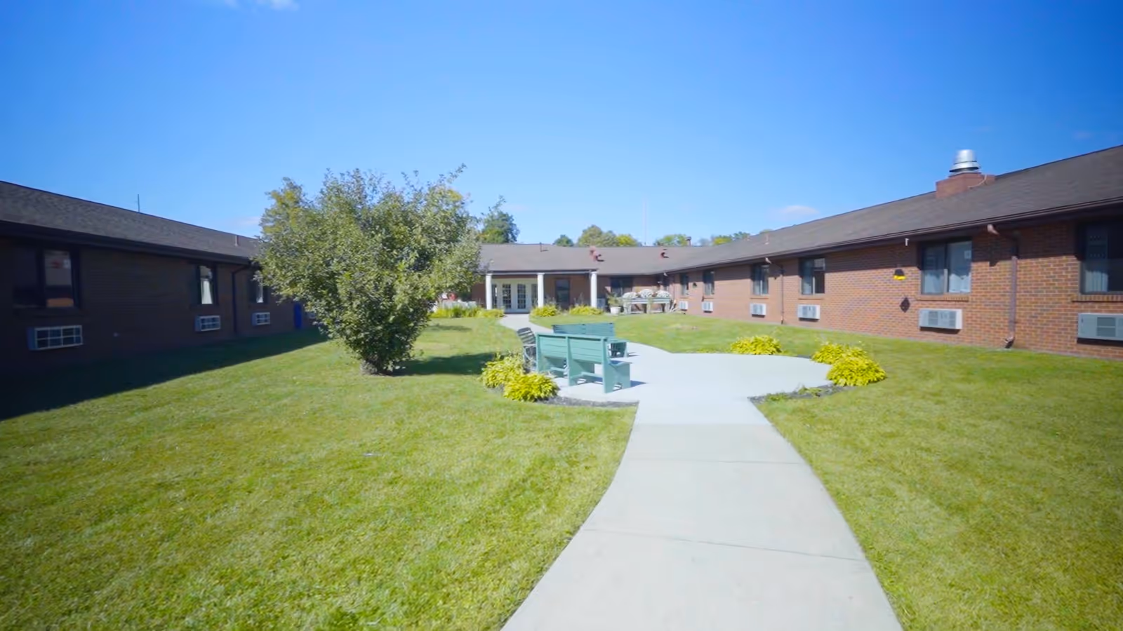 Central courtyard of a brick senior living facility with a paved walkway, benches, lawn, and single-story buildings under a blue sky.