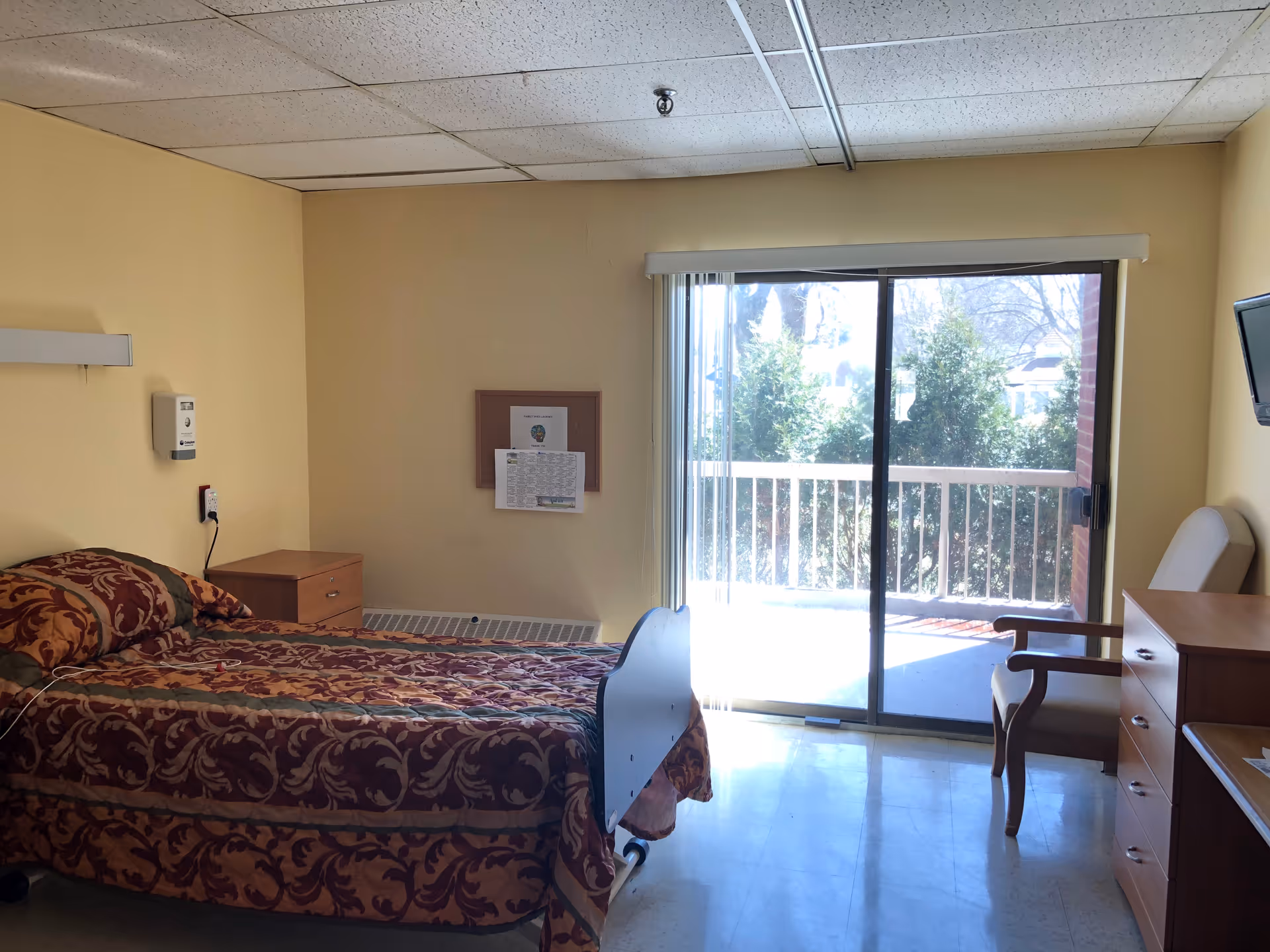 A simple, clean bedroom in a healthcare facility with a single bed covered in a patterned bedspread, a wooden nightstand, a chair, a dresser, and a wall-mounted TV. The room has a large sliding glass door leading to an outdoor balcony with greenery visible outside.