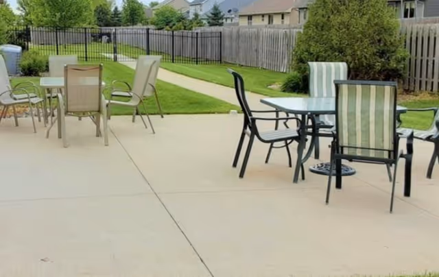 Outdoor concrete patio with glass-top tables and metal chairs beside a grassy yard and wooden fence with houses in the background.