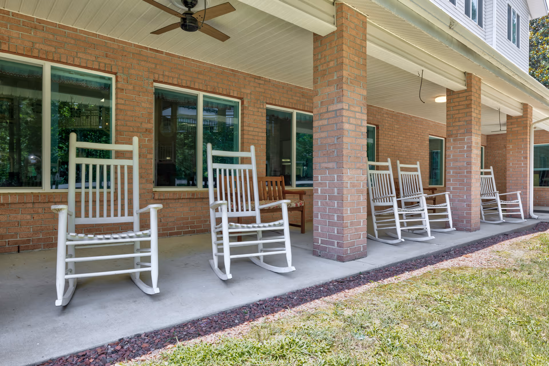 A covered outdoor patio area with several white wooden rocking chairs lined up along a brick wall with windows. The patio has a ceiling fan and overlooks a grassy area.