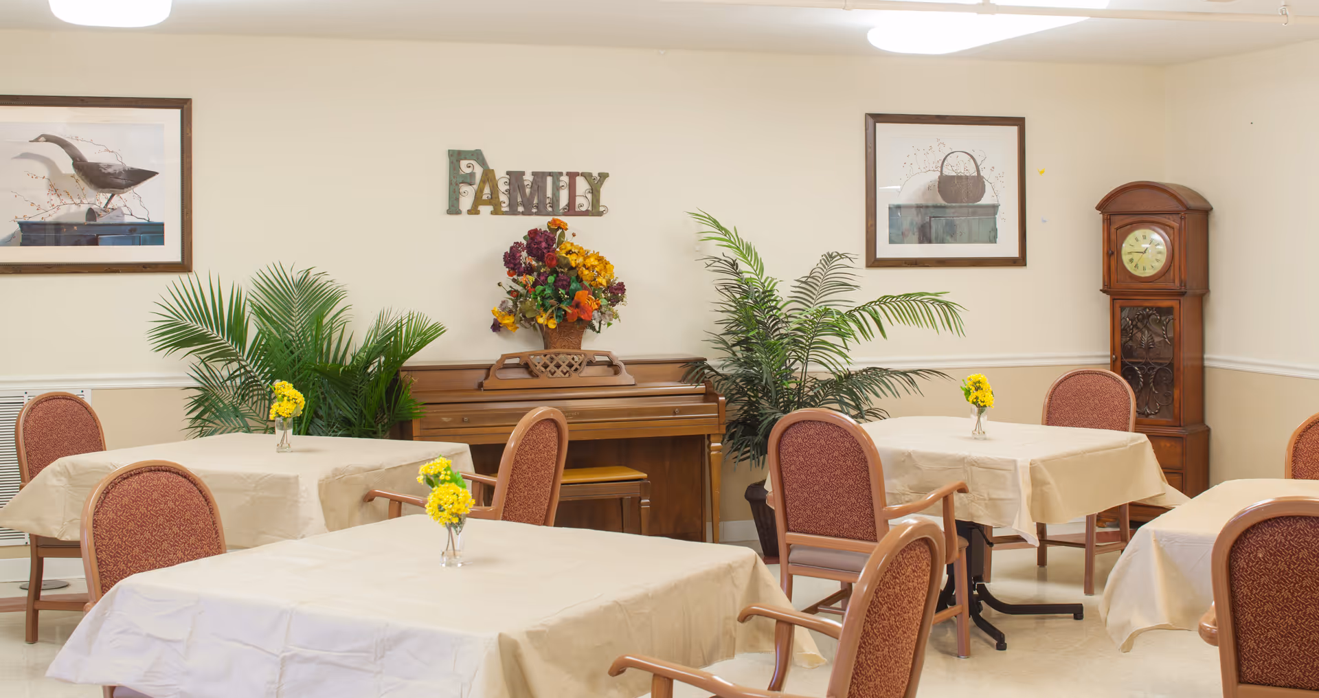 A dining room with several tables covered in beige tablecloths, each with a small vase of yellow flowers. The room features upholstered chairs with wooden arms, a wooden piano against the wall decorated with a colorful flower arrangement and a 'FAMILY' sign above it, two framed pictures, green potted plants, and a wooden grandfather clock in the corner.