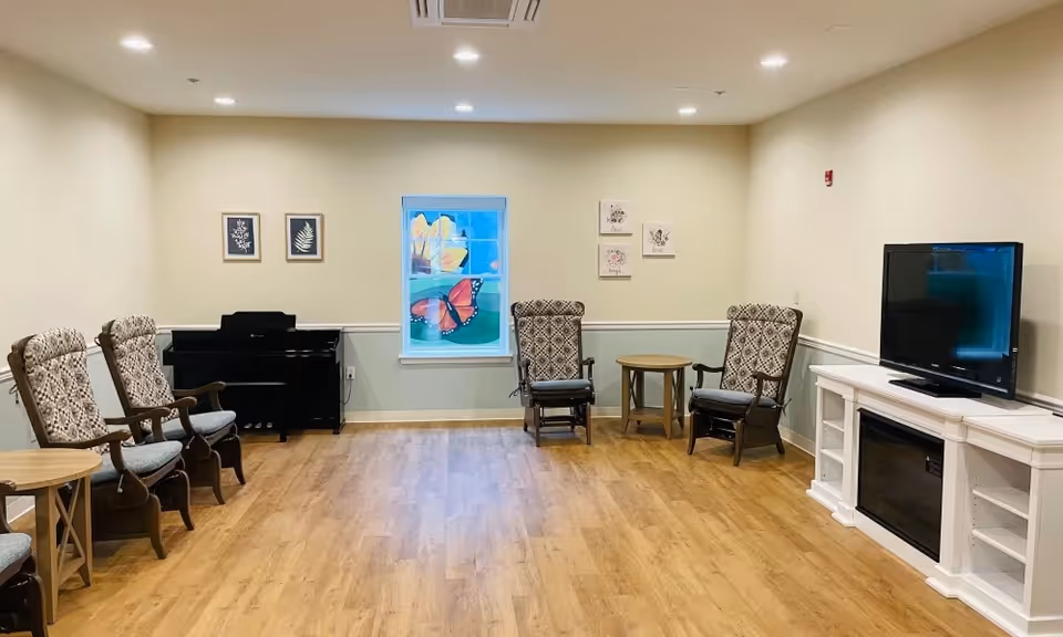 Bright common room with wood flooring, several upholstered armchairs and small tables, a piano, a TV on a white console, and a colorful butterfly window decoration.