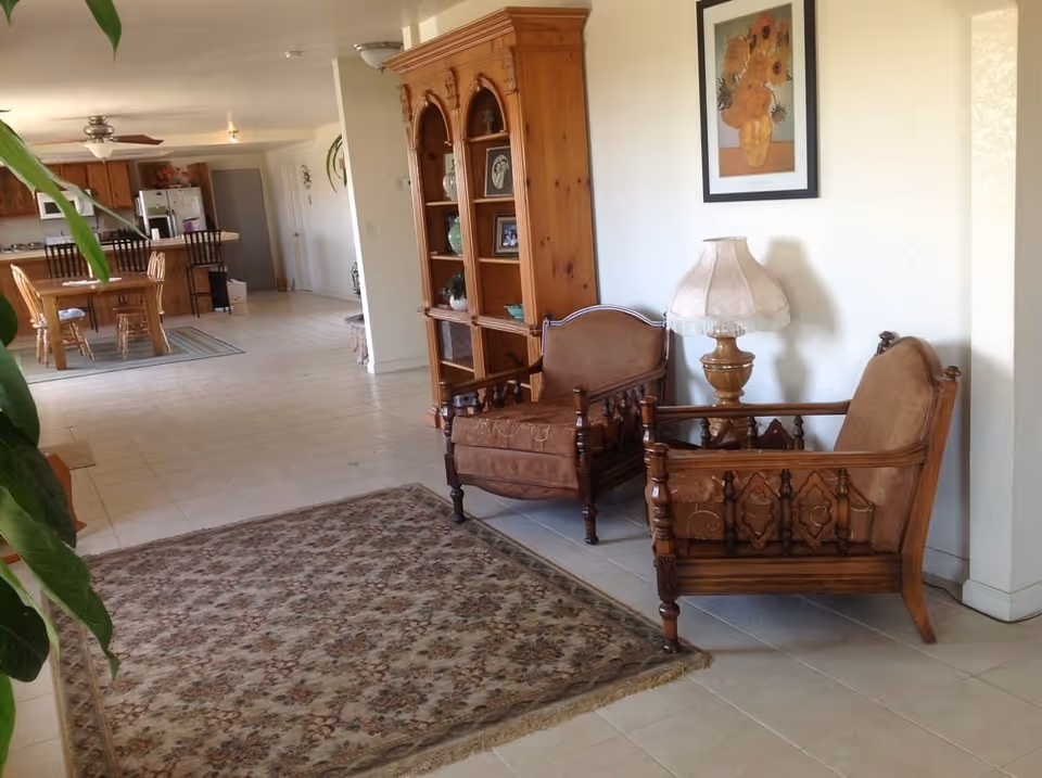 Open interior showing two carved wooden armchairs and a lamp by a bookshelf with a rug in the foreground and a dining area and kitchen visible in the background.