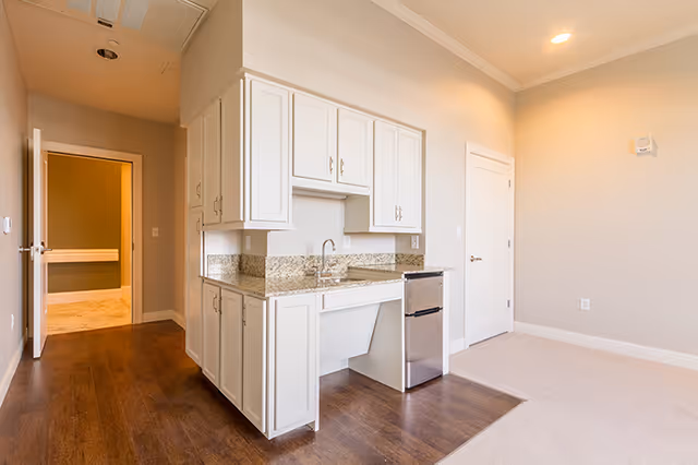 Interior view of a small kitchen area in a senior living facility with white cabinets, granite countertops, a small stainless steel refrigerator, and a sink. The kitchen area has wood flooring that transitions to carpet in the adjacent room. There is a door to the right and an open doorway leading to a hallway on the left.