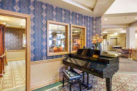 Interior view of a senior living facility featuring a black grand piano with a matching bench. The piano is placed against a wall with patterned wallpaper and two windows looking into another room. A vase with orange flowers is on top of the piano. The floor is carpeted with a decorative pattern, and the space is well-lit with ceiling lights. In the background, there is a hallway and additional seating areas.