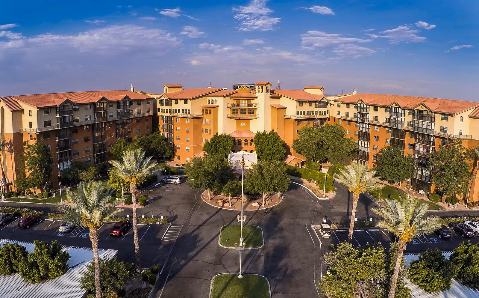 Aerial view of Casa del Sol senior living facility showing a large multi-story building with a terracotta roof surrounded by palm trees and greenery, with a parking lot in front under a partly cloudy blue sky.