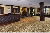 Interior view of a reception or lobby area with a dark wood reception desk on the left, patterned carpet flooring, dark wood columns, a fireplace with a painting above it, and large windows allowing natural light to enter.