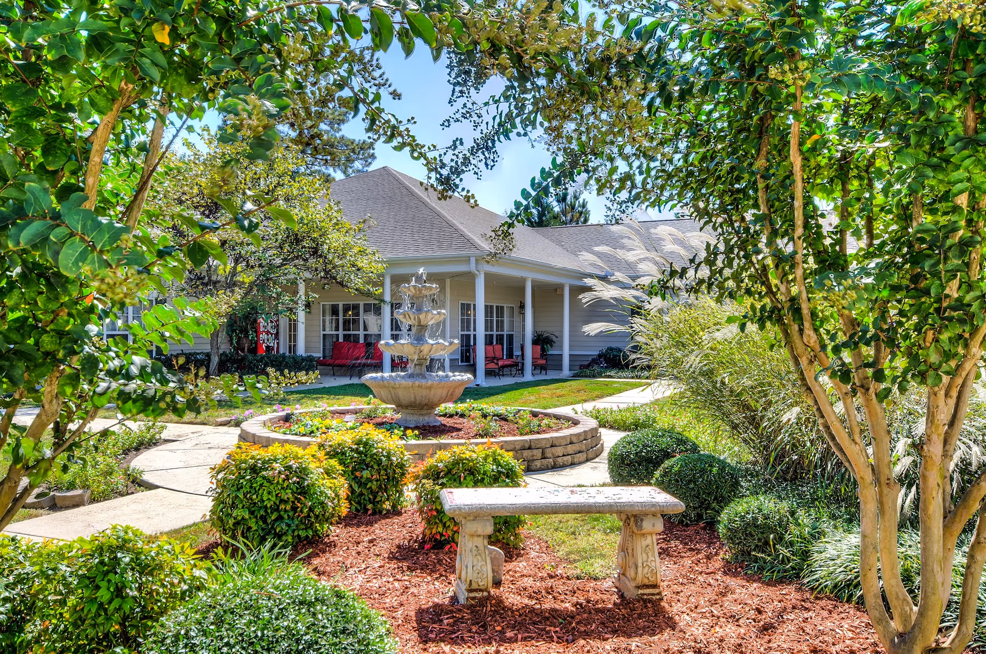 A peaceful outdoor garden area at a senior living facility featuring a stone bench in the foreground, a tiered water fountain surrounded by colorful flowers and shrubs, and a building with a covered porch and red chairs in the background. The scene is framed by green trees and bushes under a clear blue sky.