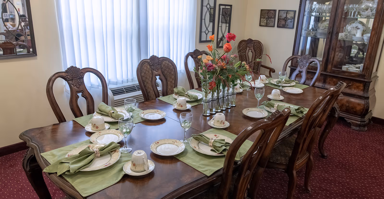 Formal dining room with a long wooden table set for eight with china, green napkins, a floral centerpiece, wooden chairs, and a china cabinet.
