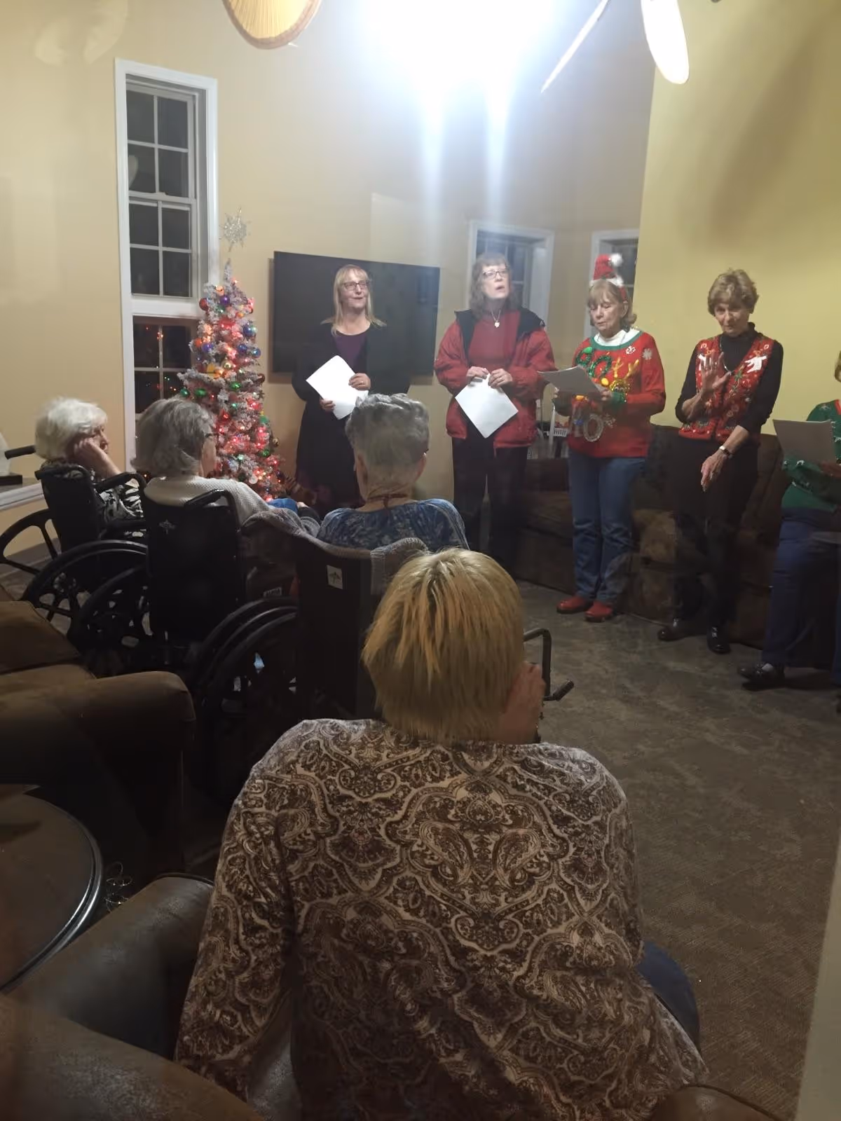 Residents seated in a living room watching a small group perform carols next to a decorated Christmas tree.