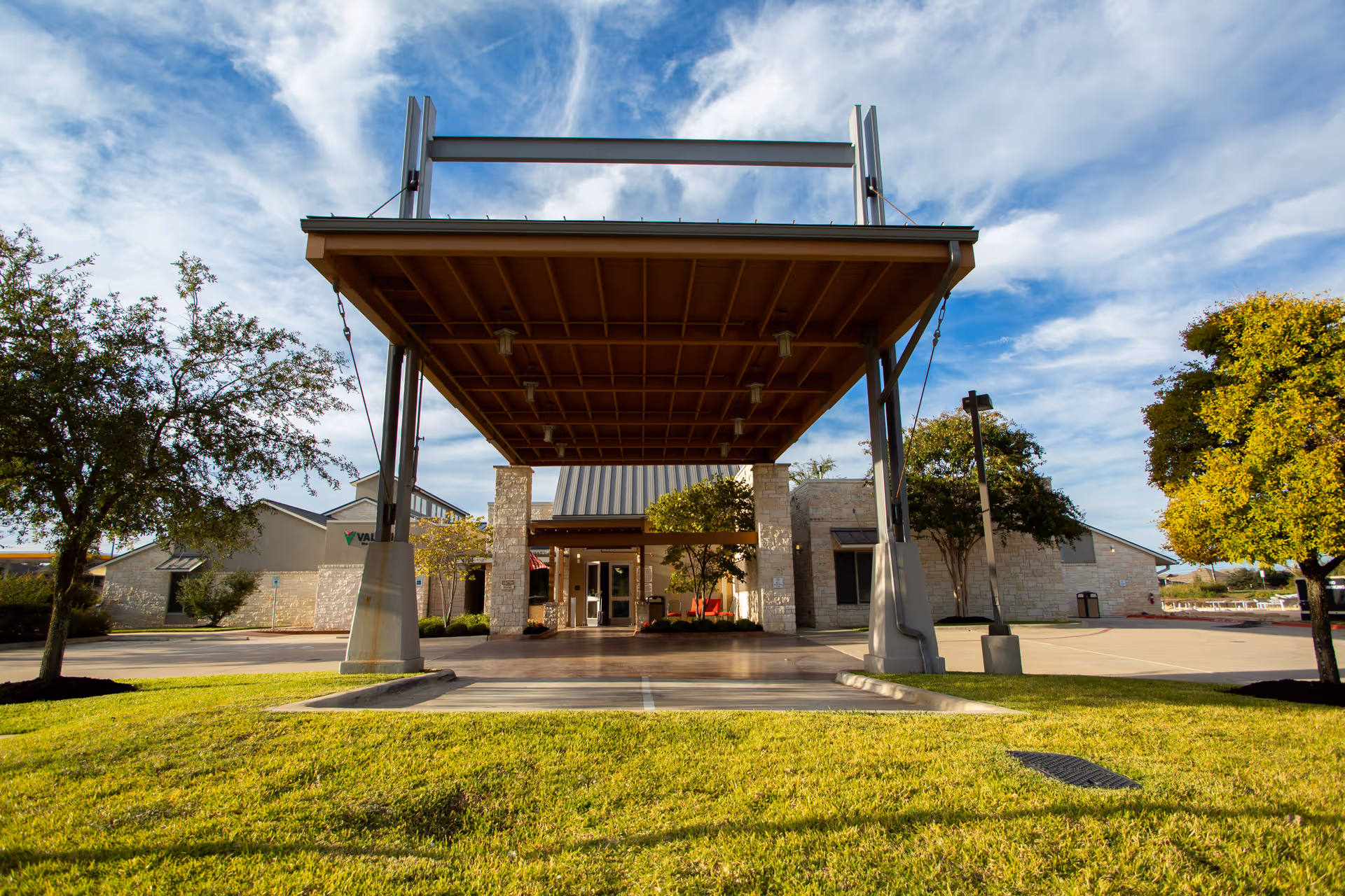 Entrance of a senior living facility with a large covered drop-off area supported by metal beams. The building is made of light-colored stone and surrounded by green grass and trees under a partly cloudy blue sky.