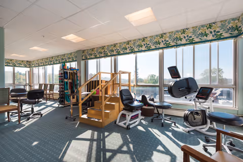 Sunlit interior activity and exercise room with large windows, seating, exercise equipment, and a small wooden therapy staircase.