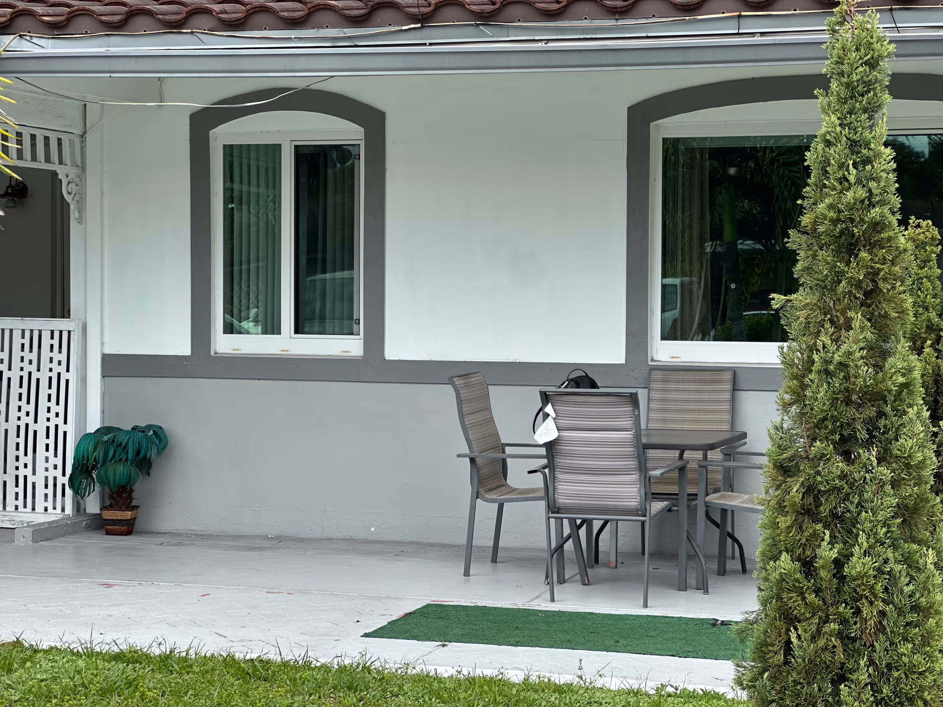 Front porch of a building with a patio table and chairs, two windows, potted plants and a small evergreen in front.