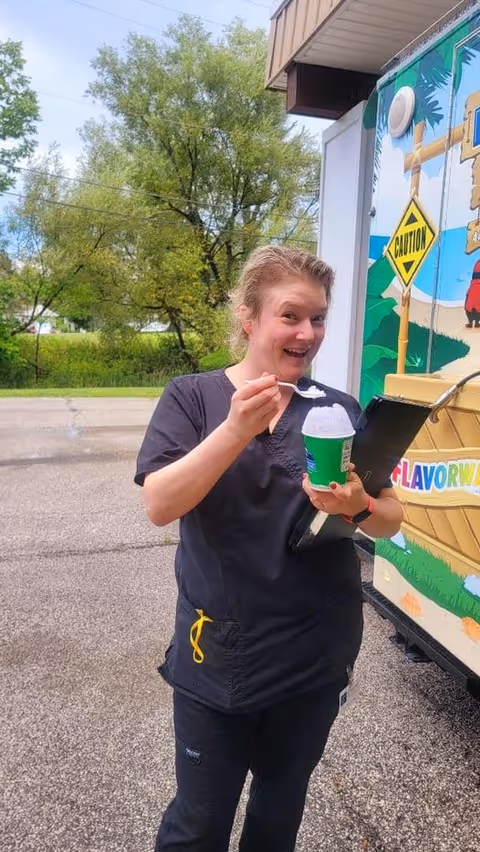 A woman wearing black scrubs is standing outside near a colorful food truck. She is holding a green cup with a white frozen treat and a spoon, smiling at the camera. Behind her, there are trees and a paved area.