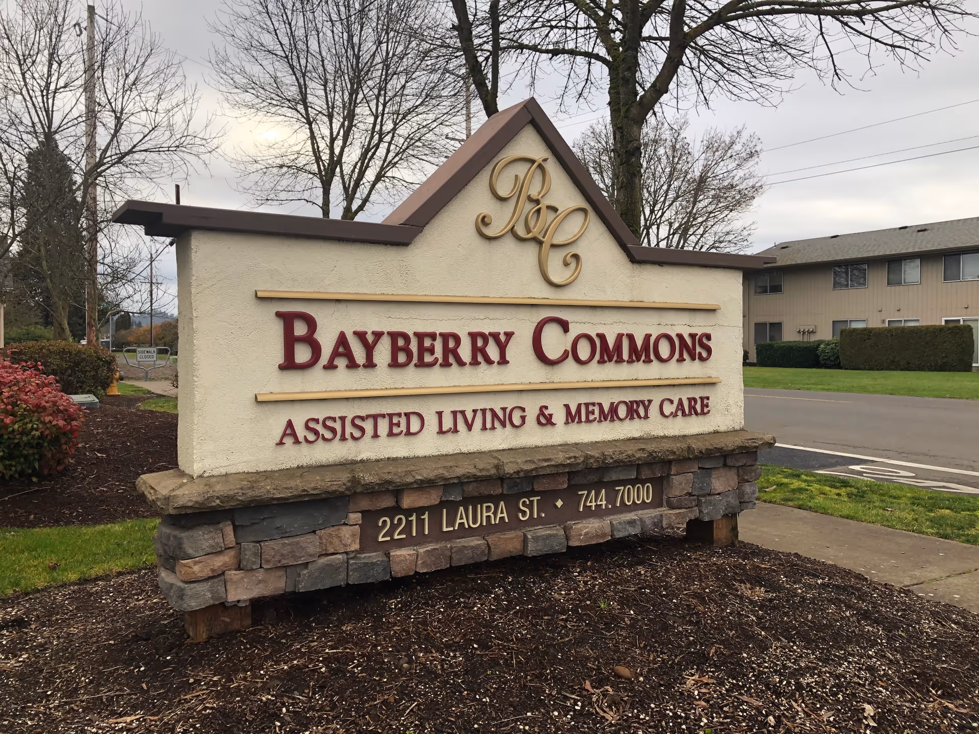 Outdoor sign for Bayberry Commons Assisted Living & Memory Care located at 2211 Laura St. The sign is made of stone and stucco with red and gold lettering. There are trees and a building in the background under a cloudy sky.