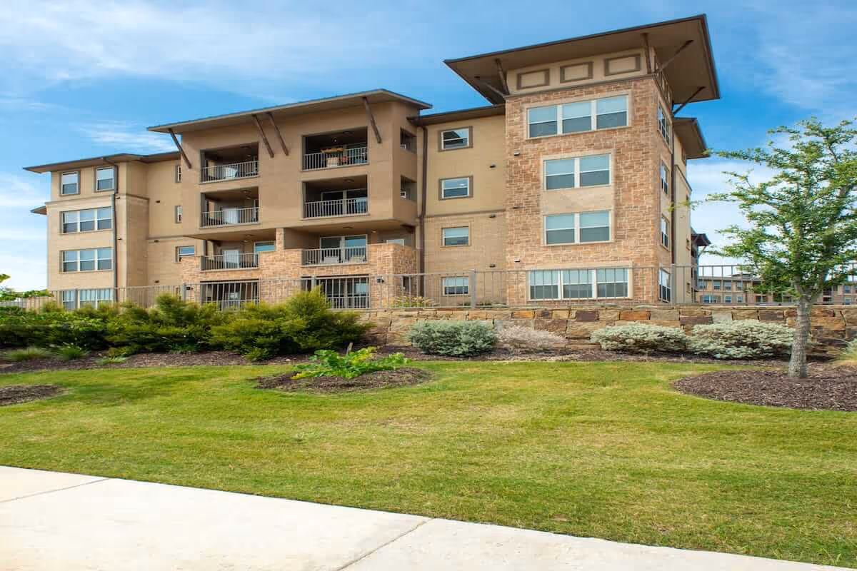 Exterior view of a multi-story senior living facility building with beige and stone facade, balconies, and large windows, surrounded by landscaped greenery and a tree under a blue sky.