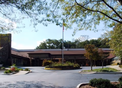 Front entrance of Meadowood with a circular driveway, flagpole, and landscaped beds in front of a low brick building.