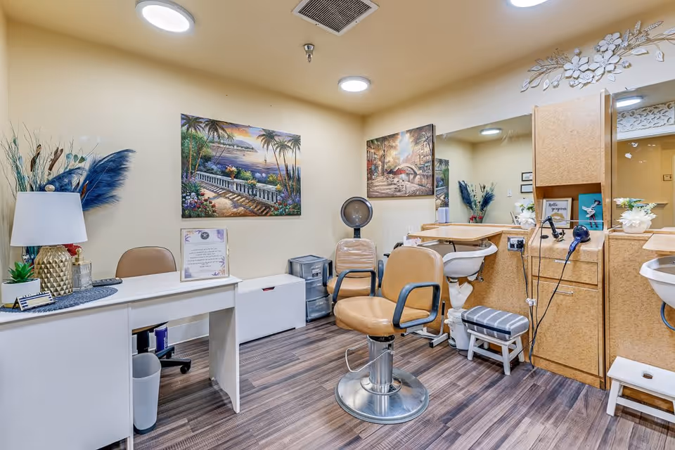 Interior of a salon area in a senior living facility with a styling chair, hair dryer, wash basin, and a desk with a lamp and decorative plants. The walls are adorned with colorful paintings and a large mirror, and the floor has wood-like flooring.