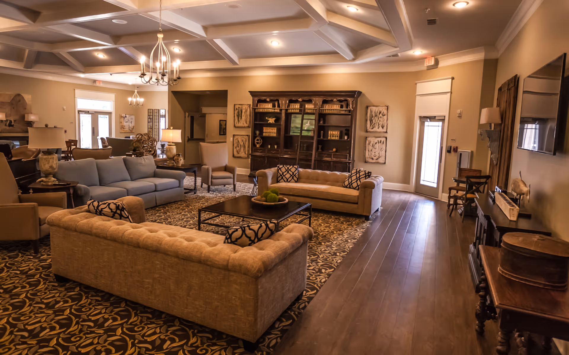 A spacious and warmly lit living room in an assisted living community featuring multiple sofas and armchairs arranged around a central coffee table on a patterned rug. The room has wooden flooring, a large wooden cabinet with decorative items, wall art, and a flat-screen TV mounted on the wall. There are several lamps and chandeliers providing ambient lighting, and a door leading outside is visible in the background.