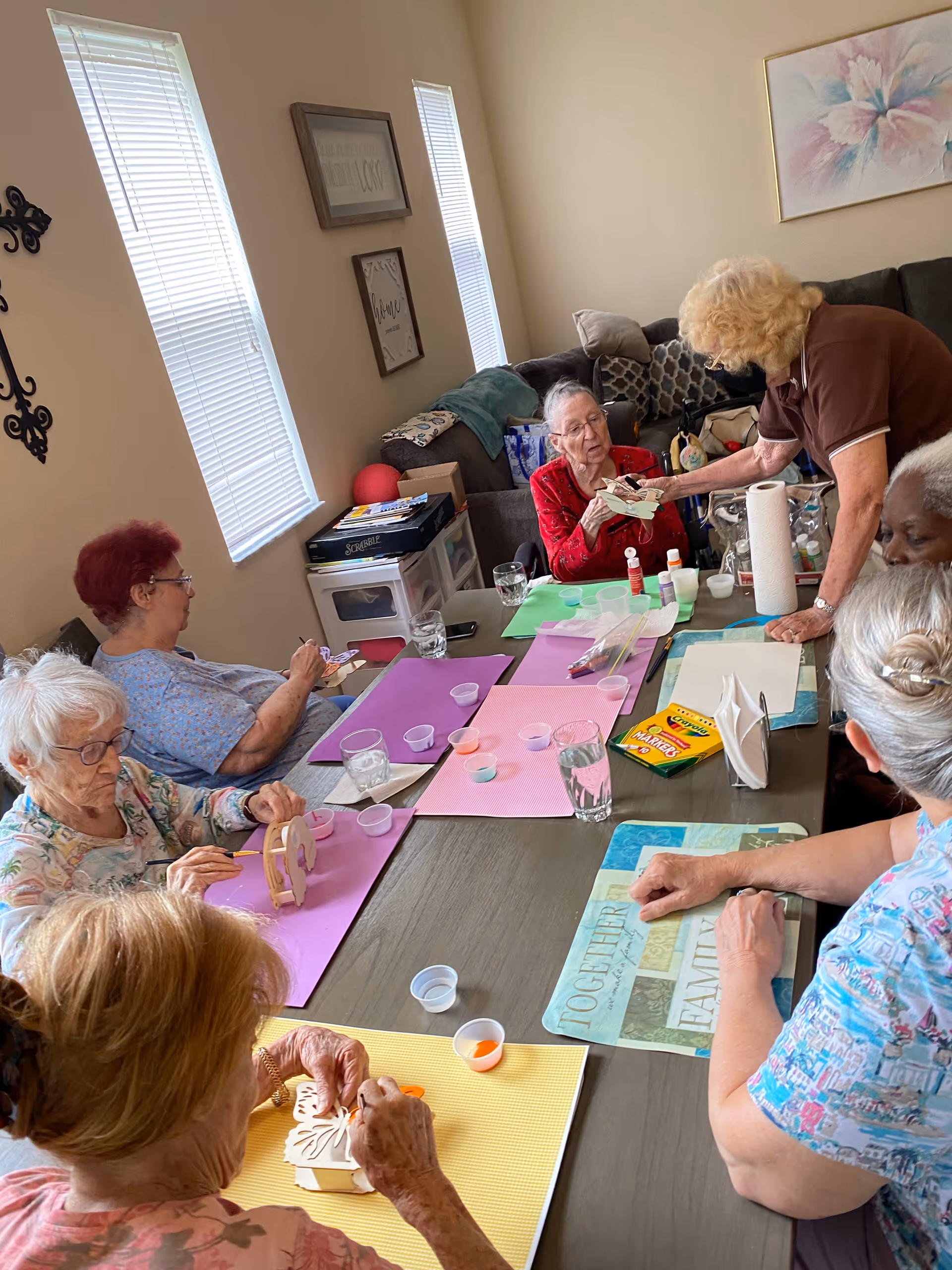A group of elderly women seated around a table engaged in a craft activity with paint and wooden objects in a well-lit room with windows and wall decorations.