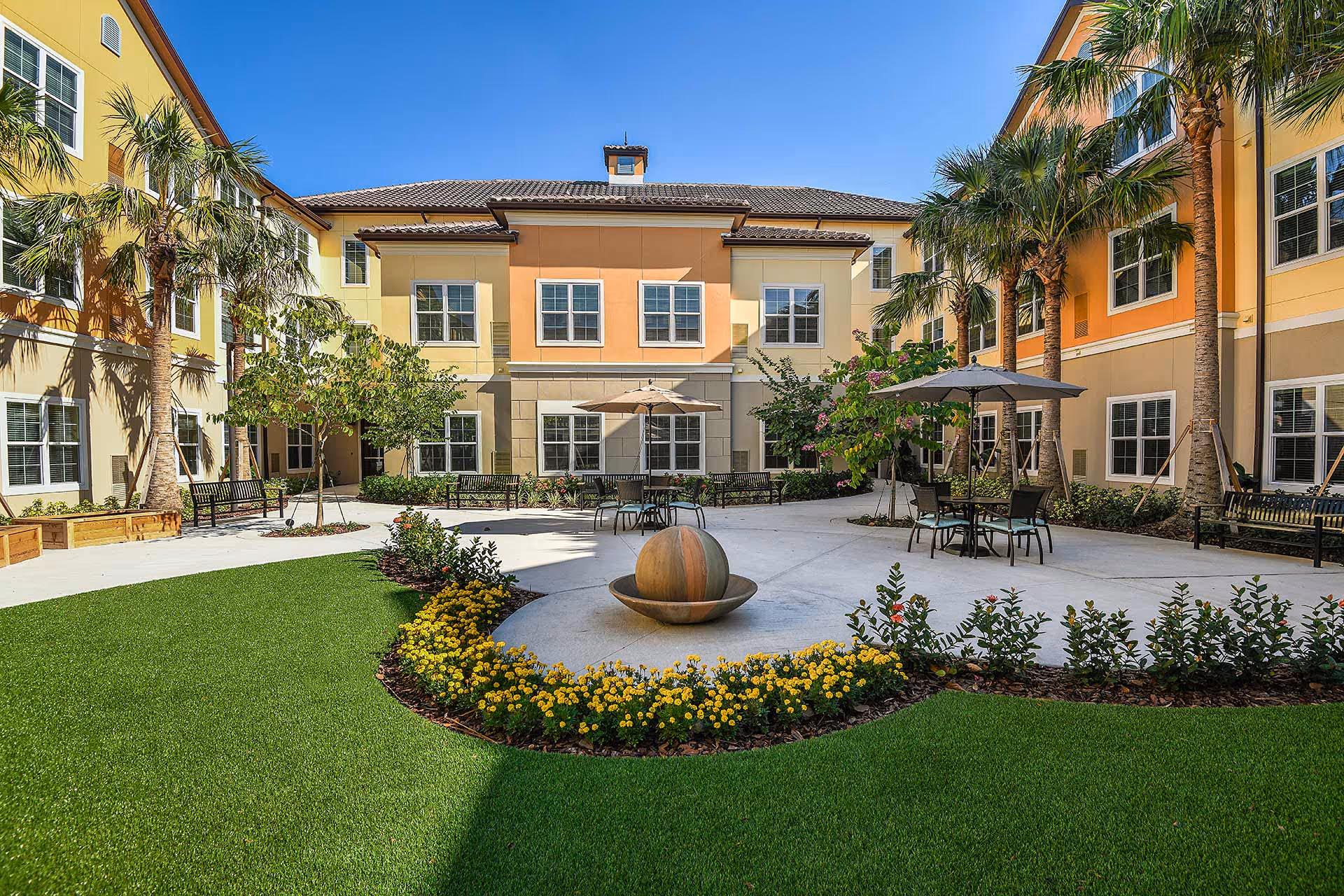 Sunny landscaped courtyard with seating, umbrellas, palm trees and a central spherical water feature in front of a three-story building.