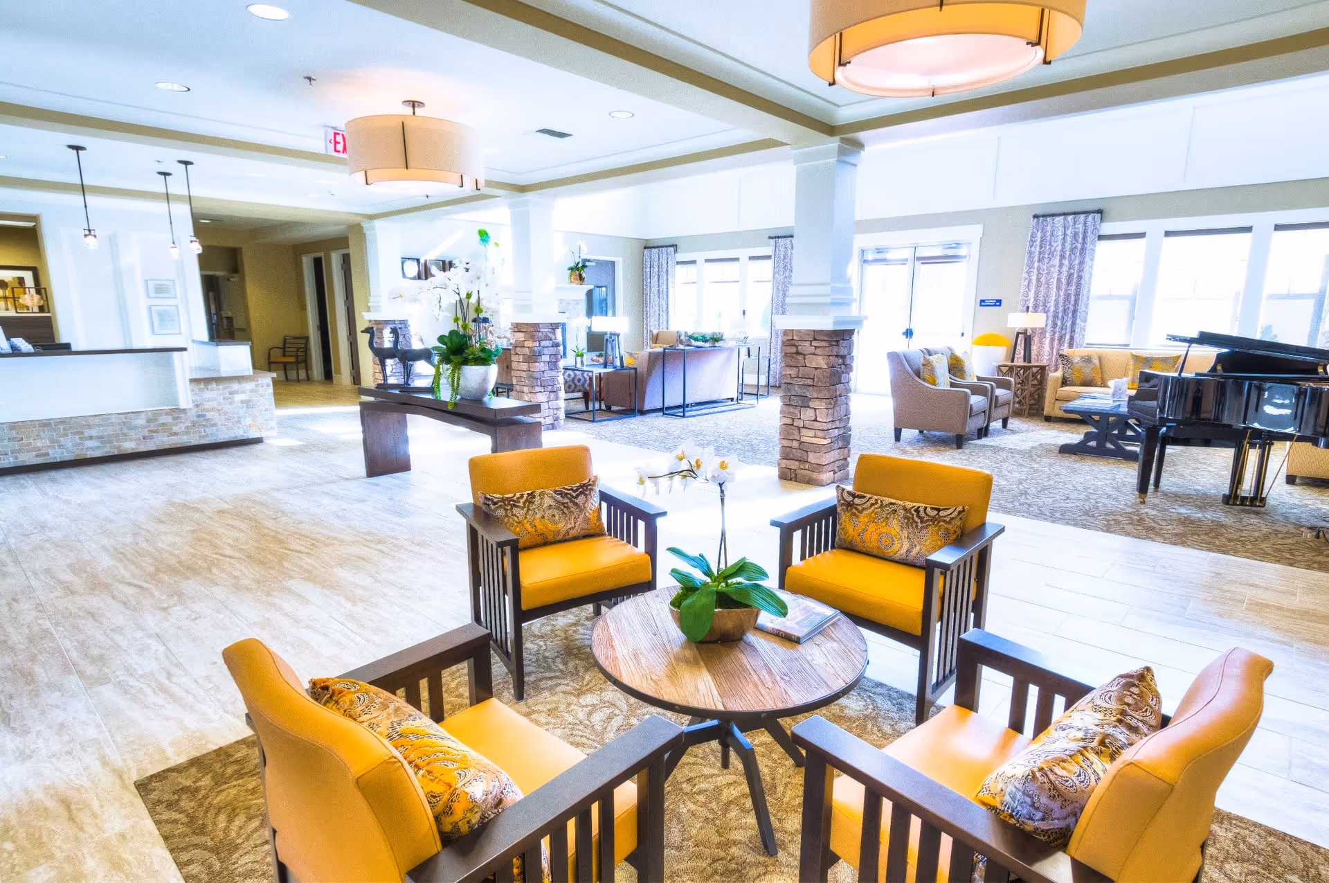Bright and spacious living room area in Carmel Village Memory Care featuring four yellow armchairs with patterned pillows arranged around a round wooden coffee table with a potted plant. The room has large windows with curtains, stone pillars, a grand piano, additional seating areas, and modern light fixtures.