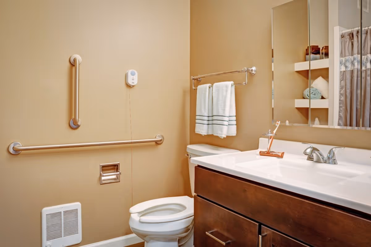 A clean bathroom with beige walls featuring a toilet with grab bars on the wall for support, a white sink with a wooden cabinet underneath, a mirror above the sink, a towel rack with two white towels, and shelves with rolled towels and decorative items. There is also a shower curtain partially visible on the right side.