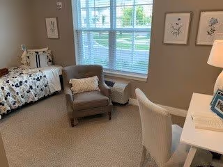 A cozy senior living bedroom featuring a single bed with floral bedding, a cushioned armchair with a decorative pillow, a white desk with a chair, a lamp, and framed artwork on the walls. A window with blinds allows natural light into the room.