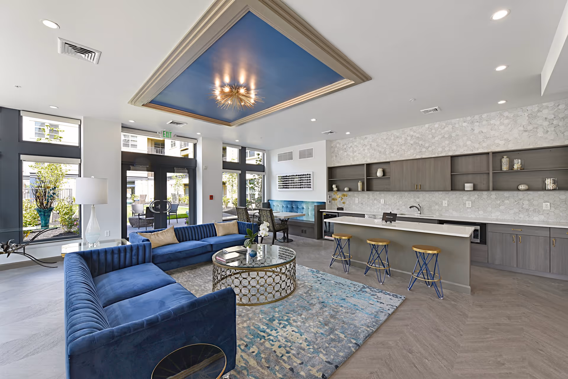 Modern senior living facility common area with two blue velvet sofas arranged around a round glass coffee table on a patterned rug. The room features a kitchen island with three blue and gold bar stools, open shelving, and a decorative backsplash. Large windows and glass doors provide natural light and a view of an outdoor patio area. The ceiling has a recessed blue panel with a stylish chandelier.