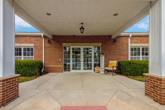 Covered entrance to a brick building with double glass doors. There are two white columns supporting the roof over the entrance. A chair with a yellow cushion and a vertical welcome sign are placed near the doors. Green bushes flank the entrance on both sides.