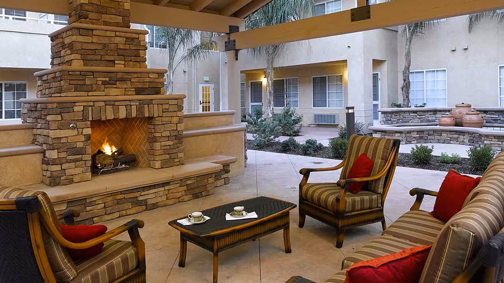 Outdoor covered seating area with a stone fireplace, cushioned chairs and sofa with red pillows, a coffee table with two cups and saucers, surrounded by a courtyard with plants and palm trees.