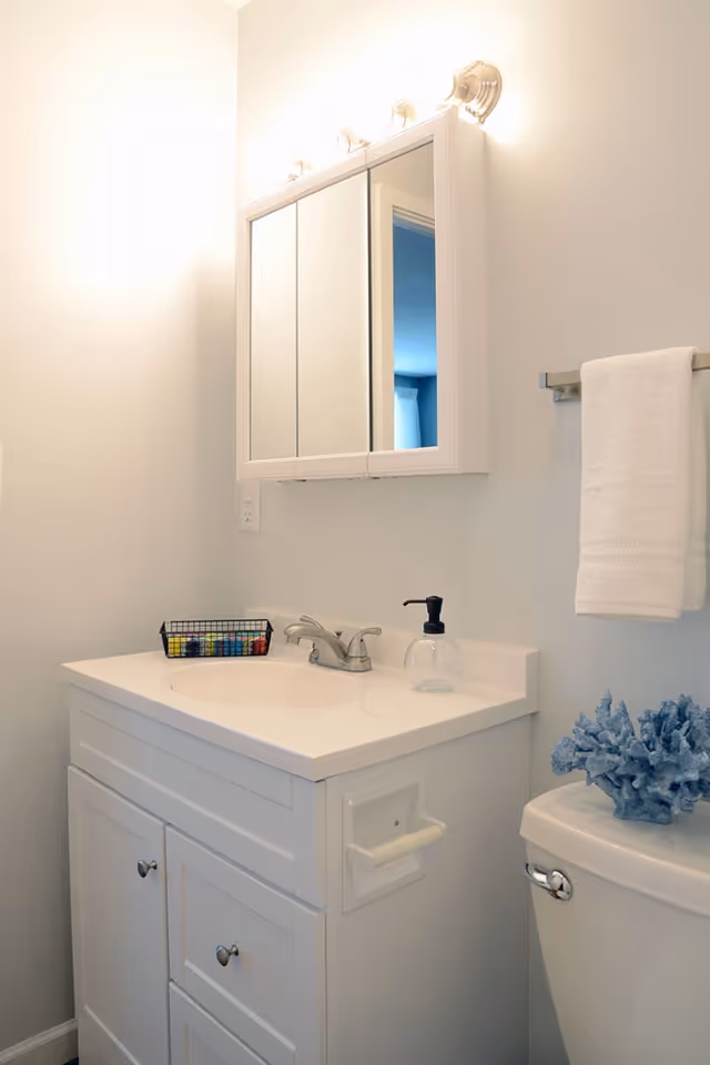 A clean and bright bathroom featuring a white vanity with a sink, a silver faucet, a soap dispenser, and a small basket with toiletries. Above the vanity is a mirrored medicine cabinet with three panels and three light fixtures on top. To the right, there is a white towel hanging on a silver towel rack and a toilet with a decorative blue coral piece on the tank lid.