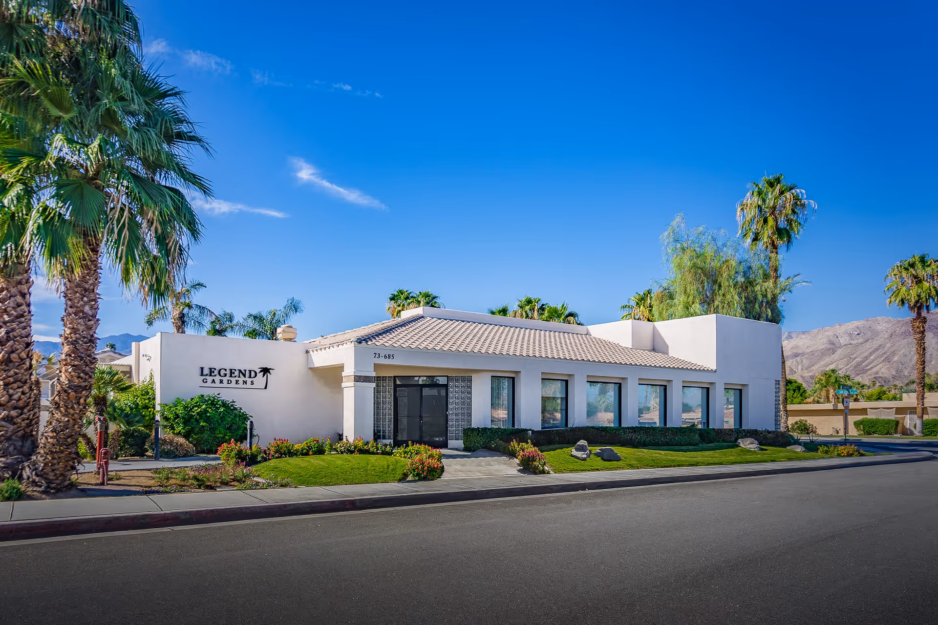Exterior view of Legend Gardens facility with a white building, tiled roof, palm trees, and landscaped greenery under a clear blue sky.
