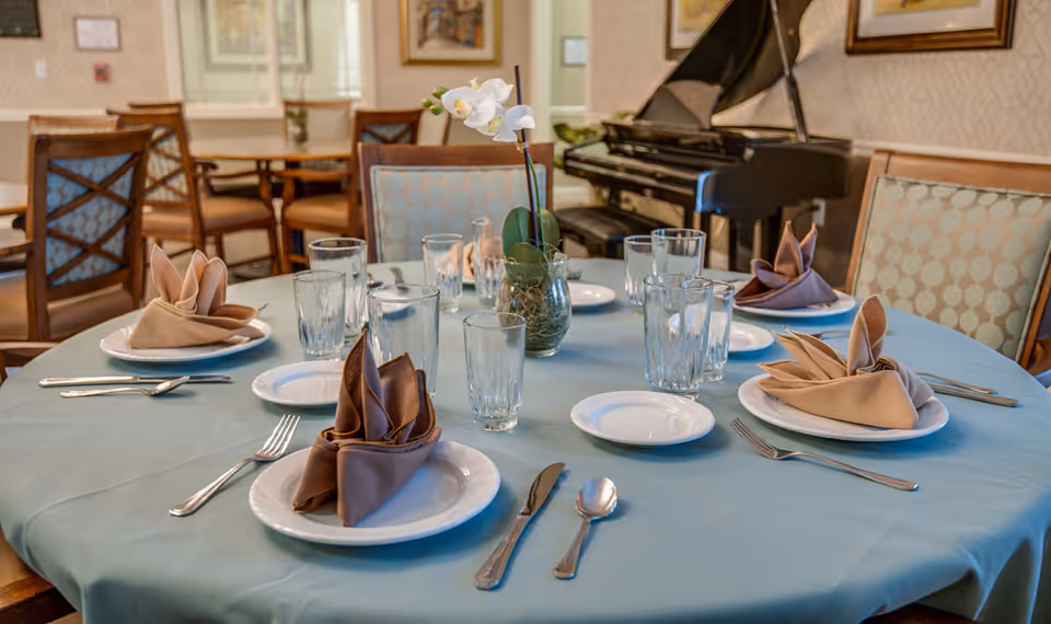 A round dining table set with plates, glasses, silverware, and folded cloth napkins in a senior living facility dining room. A small vase with white orchids is in the center of the table. In the background, there are additional tables and chairs, and a black grand piano against the wall.
