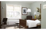 Well-lit bedroom with a single wooden bed, an armchair, a window, and bedside furnishings.
