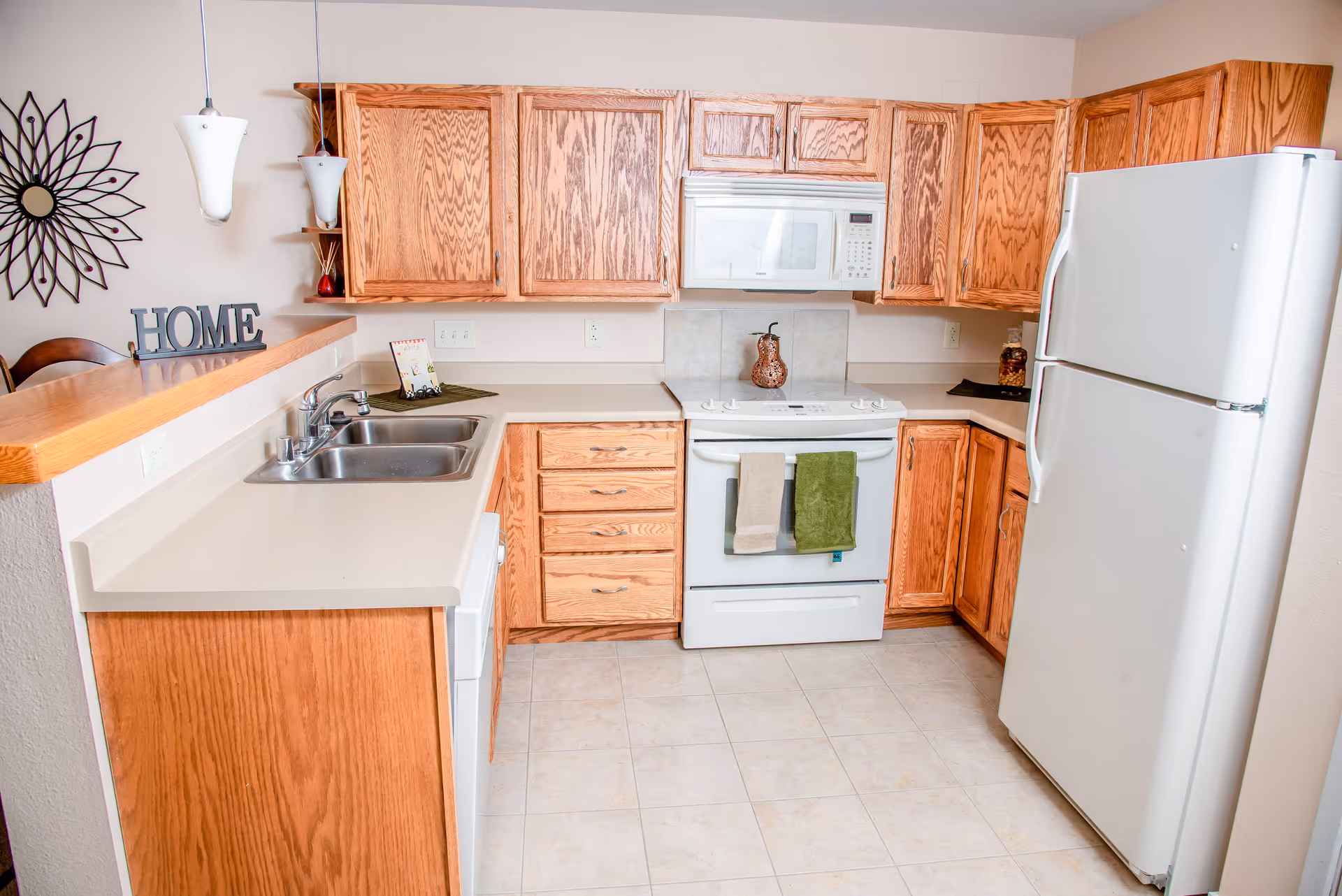Bright kitchen with oak cabinets, white refrigerator, stove and microwave, a double sink, and tiled floor.