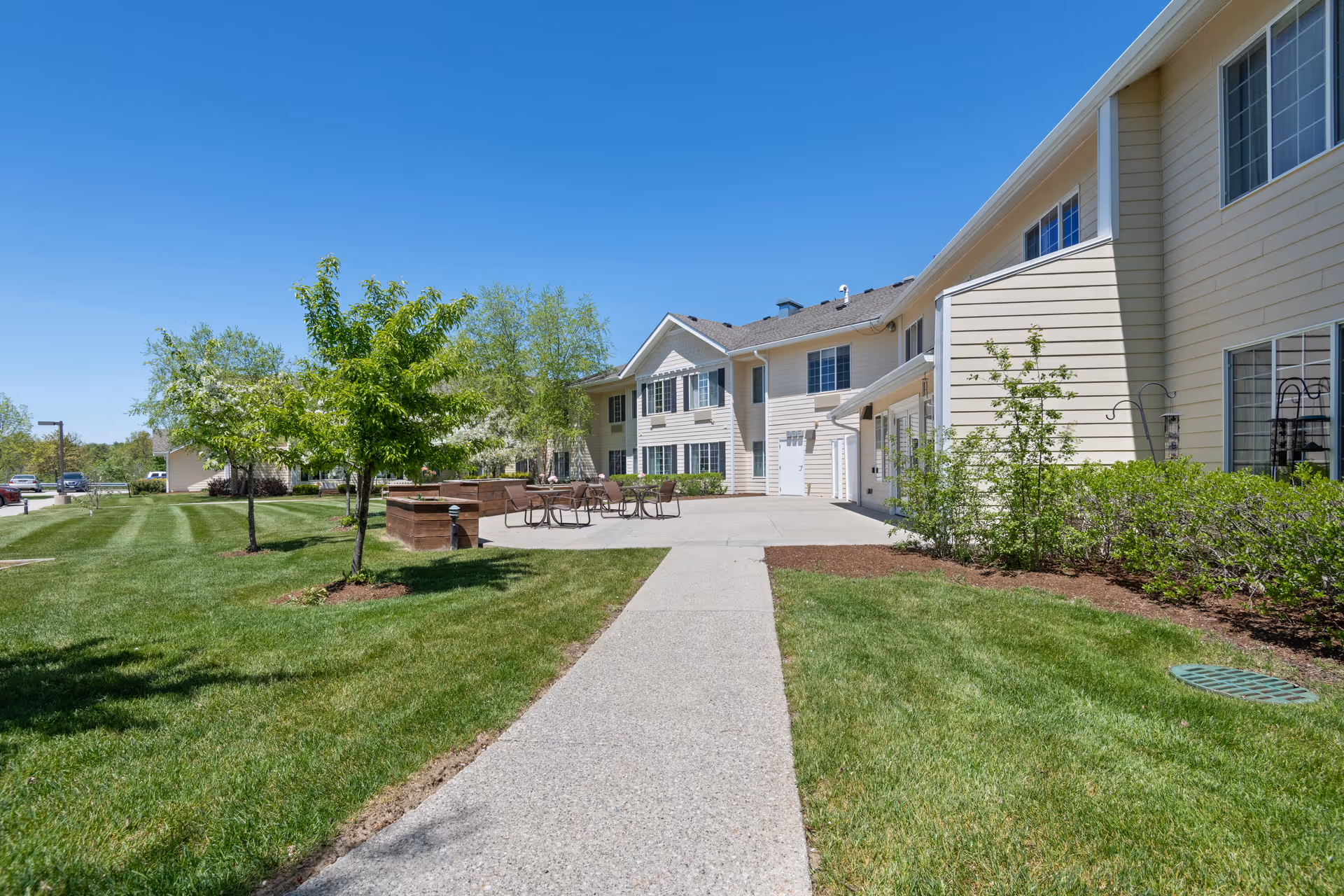 Outdoor view of a senior living facility with a paved walkway leading to a patio area with tables and chairs. The building is light-colored with multiple windows, surrounded by green grass, small trees, and shrubs under a clear blue sky.