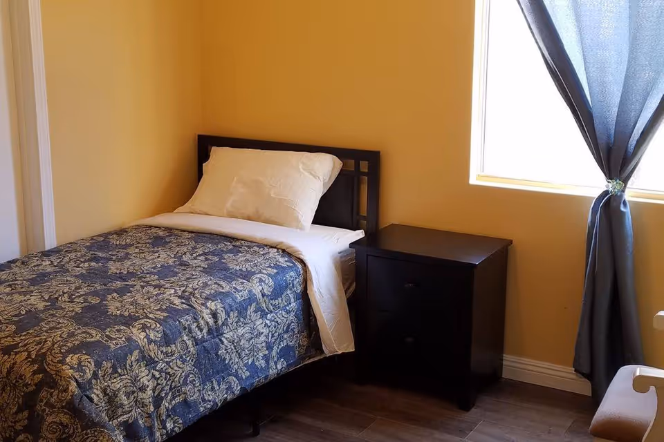 A small bedroom with a single bed covered with a blue and beige patterned comforter and a white pillow. Next to the bed is a dark wooden nightstand with two drawers. A window with dark curtains tied back allows natural light into the room. The walls are painted a warm beige color and the floor is wooden.