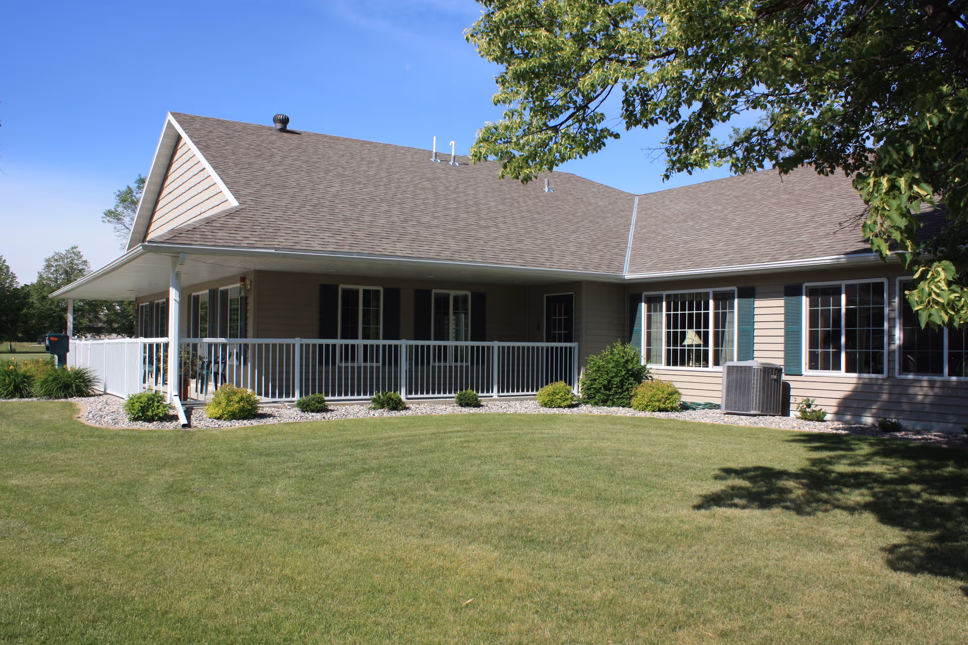 Exterior view of a single-story residential building with beige siding, a brown shingled roof, and white railing around a porch. The building is surrounded by a well-maintained lawn and some bushes, with a tree partially visible on the right side under a clear blue sky.