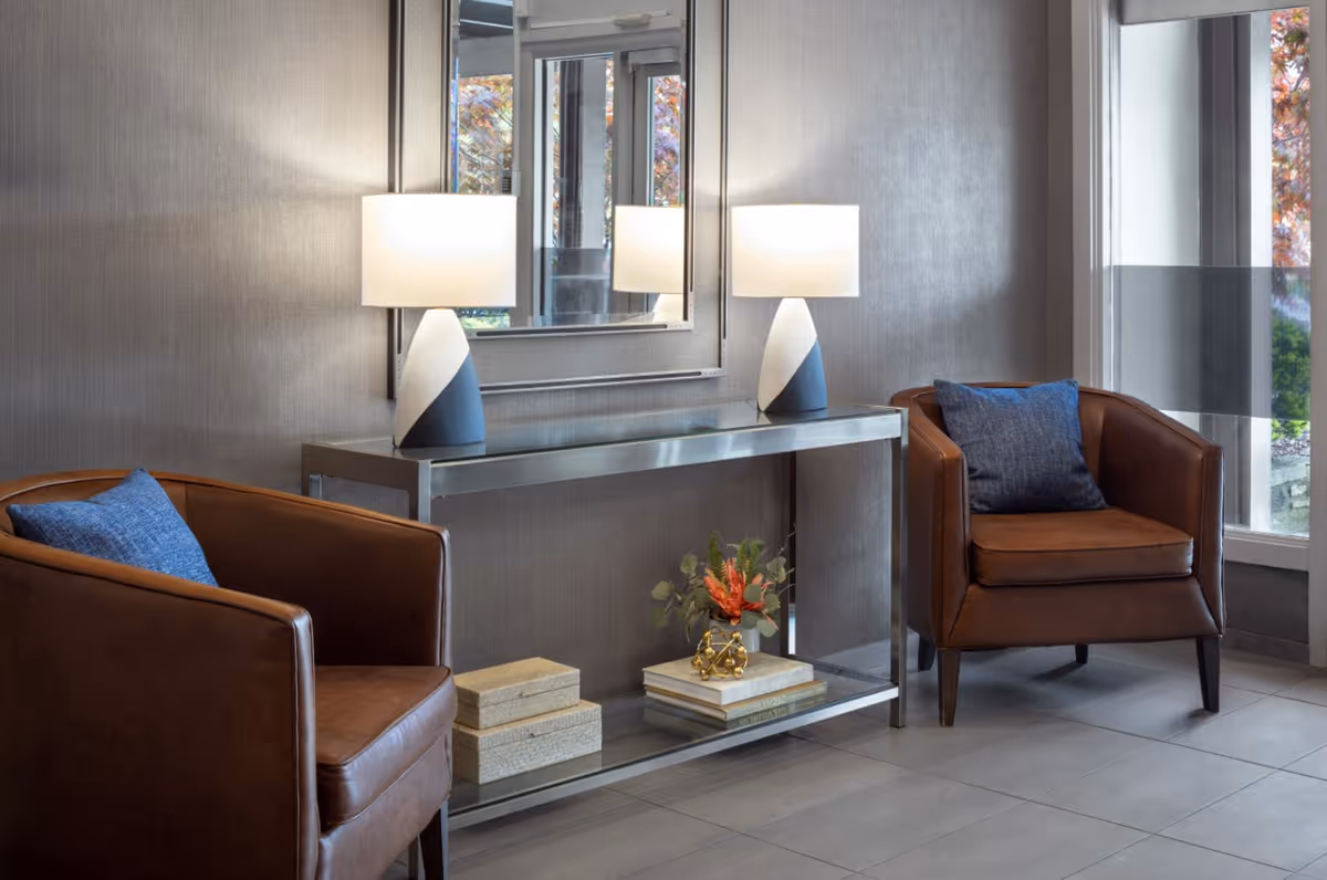 Two brown leather armchairs flank a console table with two lamps and a mirror in a modern lobby seating area.